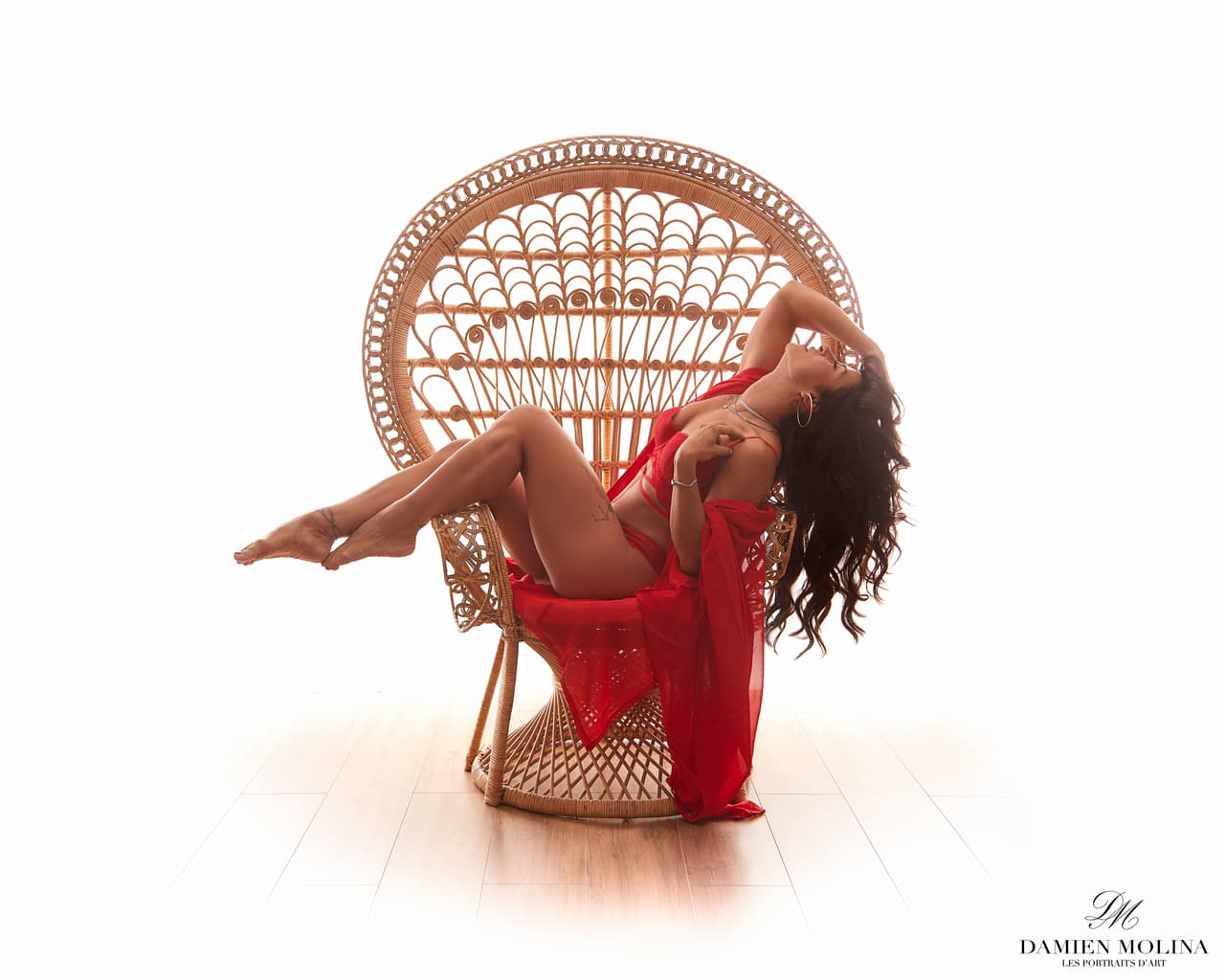 Passion en rouge : Séance boudoir glamour à Strasbourg - Damien Molina - Photographe Photo boudoir d'une femme en lingerie rouge assise sur un fauteuil en osier de type Emmanuelle, capturée sur un fond lumineux pour une séance sensuelle à Strasbourg.