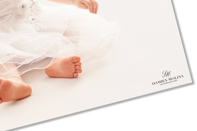 Signer les photos 10w - Damien Molina - Photographe Close-up of a baby's bare feet peeking from a flowing white dress on a pale backdrop, with a Damien Molina watermark in the corner.