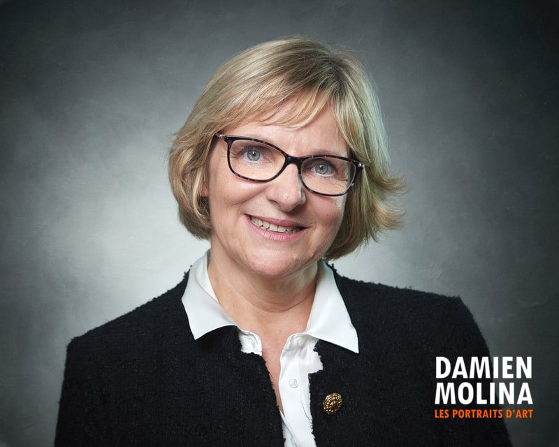 Signer les photos 5 - Damien Molina - Photographe Smiling middle-aged woman with short blond hair and glasses, wearing a white blouse and black blazer in a studio portrait.